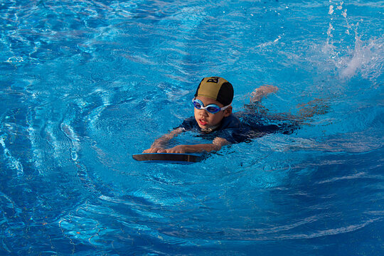 6 years old young boy who a swimmer wearing goggle with dark blue suit and yellow striped black cap. Lonely child warm up with swimming kickboard before practicing to swim in the blue swimming pools.