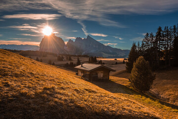 Alpe di Siusi, Italy - Autumn sunrise with a wooden chalet at Seiser Alm in South Tyrol in the Dolomites mountain range with Saslonch (Sassolungo or Langkofel) mountain and rising sun at background