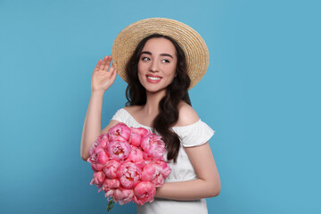 Beautiful young woman in straw hat with bouquet of pink peonies against light blue background