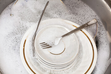 Washing dishes. Forks, spoons, plates of different sizes in the sink under running water. An image about housework, apartment care and cleanliness.