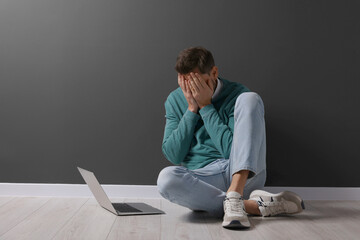 Upset man sitting on floor near laptop against black wall. Space for text
