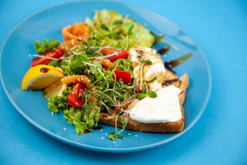 Healthy breakfast. Avocado and salmon, poached egg and toast, salad and vegetables. Microgreens on a plate. Food bowl. Lunch on a blue plate. Sauce and sesame seeds, lemon tomatoes. Top view.