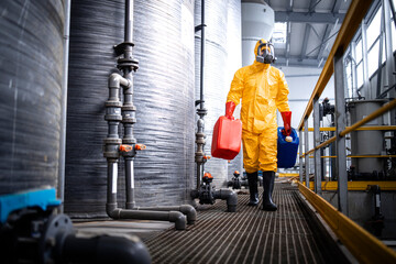 Factory worker in protective equipment walking by acid storage tanks and carrying aggressive and...