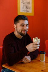 A man is sitting and smiling in a cafe and holding a shawarma in his hand.
