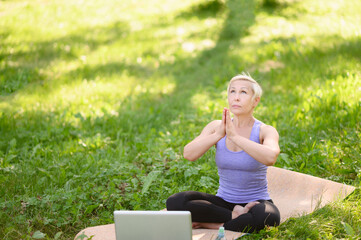 Athletic middle aged woman doing yoga with laptop online outdoors in park sitting in lotus position .Concept of online training, stretching, yoga.