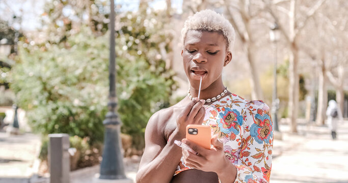 Transgender woman applying herself lipstick looking at mobile phone while standing outdoors. - Powered by Adobe