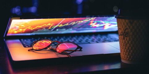 A laptop half closed in the dark with colourful glow, cup of coffee and glasses.