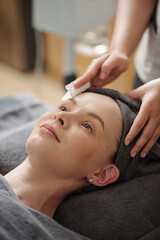 Naklejka premium Cosmetologist wiping face of young woman with cotton pad soaked in micellar water