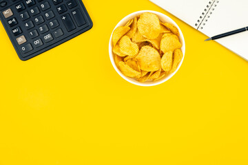 A plate of potato chips, a calculator and a notebook on a yellow background.