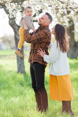 Happy young family with little son walks in blooming garden. Mom and dad with 1 year old baby. Spring.