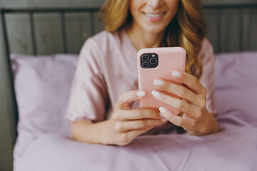 Cropped young woman wear t-shirt pajama sits on bed hold in hand use mobile cell phone rest relax spend time in bedroom lounge home in own room wake up dream be lost in good mood day. Focus on device.