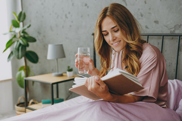 Young woman wear purple t-shirt pajama sit in bed read book drink water rest relax spend time in bedroom lounge home in hotel room wake up dream be lost in reverie good mood day. Real estate concept.