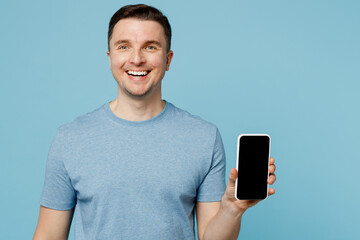 Young satisfied cheerful smiling happy caucasian man wear casual t-shirt hold in hand use mobile cell phone with blank screen workspace area isolated on plain pastel light blue cyan background studio.