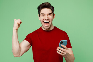 Young happy overjoyed man he wearing red t-shirt casual clothes hold in hand use mobile cell phone do winner gesture isolated on plain pastel light green background studio portrait. Lifestyle concept.