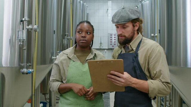 Medium shot of African American woman and Caucasian man in aprons standing next to large steel fermenter tanks at craft brewery, looking at data on clipboard and discussing beer maturing process