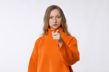 Fototapeta na wymiar It's your fault. Portrait of disappointed young woman frowning, pointing index finger at camera, being mad, blaming all her failures on you, stands in orange sweater against neutral studio background