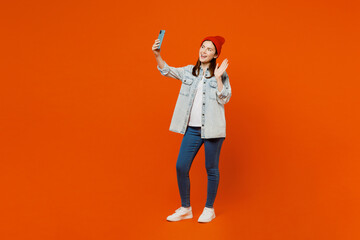 Full body young happy woman wear denim shirt white t-shirt red hat doing selfie shot on mobile cell phone post photo on social network waving hand isolated on plain orange background studio portrait
