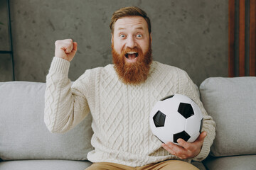 Young overjoyed happy man fan wear casual clothes cheer up support football team sit on grey sofa...