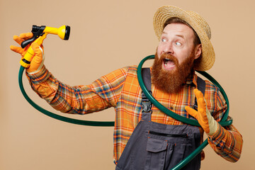 Young bearded man wear straw hat overalls work in garden play with hose with sprinkler watering...