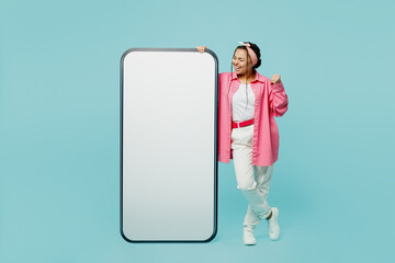 Full body young woman wear pink shirt white t-shirt headscarf big huge blank screen mobile cell phone with area do winner gesture isolated on plain pastel light blue cyan background studio portrait.