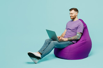 Full body smiling happy young IT man he wears purple t-shirt sit in bag chair hold use work on...