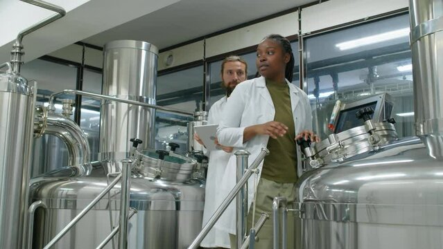 Full dollying shot of multiethnic brewery employees working together on brewhouse with large steel tanks, adjusting equipment and discussing brewing process