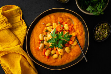 Healthy lunches, tomato and carrot puree soup, beans with pumpkin seeds, healthy tasty legumes, top view of a plate of soup on a black background