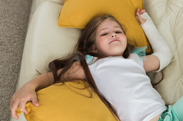 Child with a cast on a broken wrist or arm smiling and having fun on a couch. Positive attitude,...