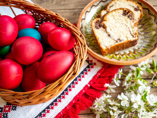 basket of red Easter eggs next to flowering branches and plate with sweet sponge cake or cozonac on wooden table