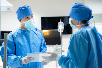 Selective focus of a young Caucasian male doctor in surgical uniform, face mask, and gloves, standing holding a clipboard with medical report while talking to female doctor in a modern operating room.