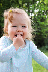 portrait of funny smiling little baby girl with finger in mouth against nature background outdoors