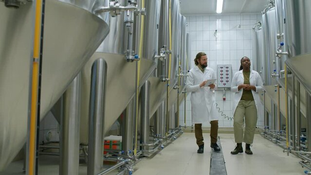 Full pan shot of fermentation facility at craft brewery, and two multiethnic male and female employees in white coats walking and pointing to bright beer tanks, engaged in lively discussion