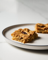 Two squares of Blondie cookies with pecan nuts on white plate. White chocolate sweet and tasty, chewy cookie bars. Close up