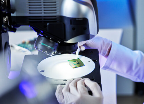Hands of technician examining electrical chip under microscope at E-Laboratory