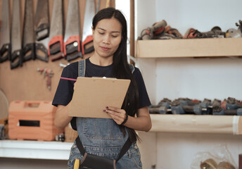 Portrait of female carpenter holding clipboard standing in carpentry workshop. Asian woman woodworker confidently designing woodwork furniture with professional skill. Small business and expertise.