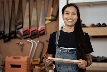 Portrait of female carpenter holding clipboard standing in carpentry workshop. Asian woman woodworker confidently designing woodwork furniture with professional skill. Small business and expertise.