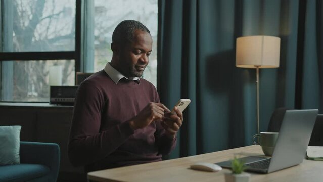 Side footage of smiling African man looking at smartphone, typing message and scrolling social media. Portrait of joyful freelancer during coffee break in comfort atmosphere of home. Indoors