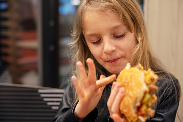 happy girl with long blond hair eats a burger with pleasure licking her fingers