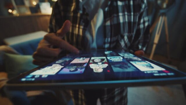 Close-up footage of hands of unrecognizable young woman in casual checkered shirt scrolling through screen of digital tablet. Girl using streaming platform for leisure and looking for movies