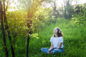 Young happy woman listening to music in headphones sitting on green grass enjoying nature