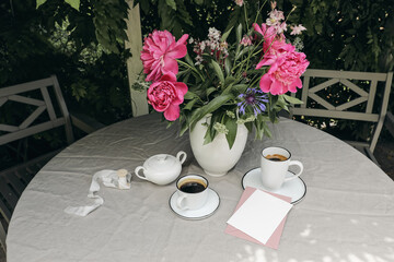 Summer breakfast on terrace. Cups of coffee, blank greeting cards and peonies bouquet. Gey linen table cloth. Outdoor dining. Dark green garden, yard or patio background. Leisure, lifestyle concept.