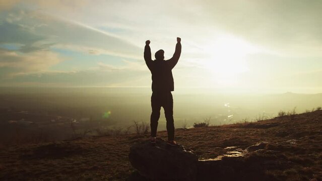 One Successful Happy Travel Man Standing Top Joy and Raises Hands Up. Joyful Silhouette Man Looking at View Sunset Sky Behind Mountains. Adventure Traveling Hike Man Hands Raised on Hill, slow motion