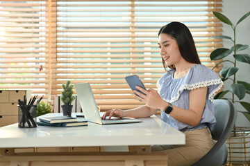 Attractive asian woman freelancer holding smart phone and using laptop at modern home office