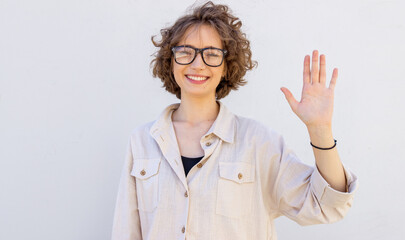 Friendly young woman in glasses smiles happily and waves her right hand in the meaning of hello, hi or goodbye. The student shows five fingers. © A Stock Studio