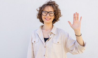 Friendly young woman in glasses smiles happily and waves her right hand in the meaning of hello, hi...