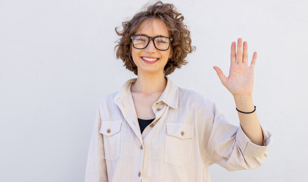 Friendly young woman in glasses smiles happily and waves her right hand in the meaning of hello, hi or goodbye. The student shows five fingers.