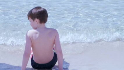 happy cool child, healthy boy 9-10 years old sits on the sand on sea, river shore, frolic, playing in water, splashing in splashes, concept fear of water, fear of drowning swimming skills