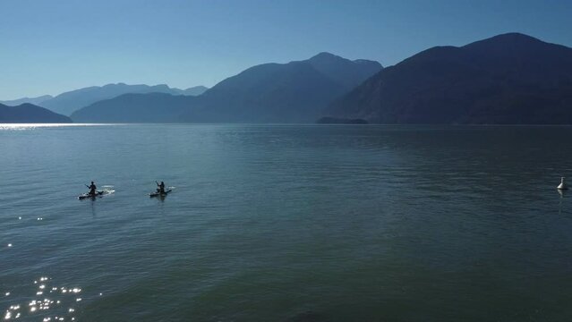two kayakers paddling back to shore in the ocean of Porto Cove, BC. blue still water breathtaking mountain scenery, abandoned industrial structure repurposed as tourist attraction adds a unique touch
