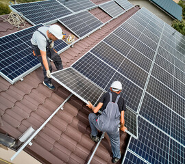 Men installers installing photovoltaic solar moduls on roof of house. Engineers in helmets building solar panel system outdoors. Concept of alternative and renewable energy. Aerial view.