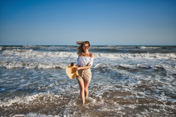 Cute beauty runs on the waves, holding a hat in her hands. Sea beach. Lifestyle girl with glasses and a hat at sea. Pastel colors of clothing.